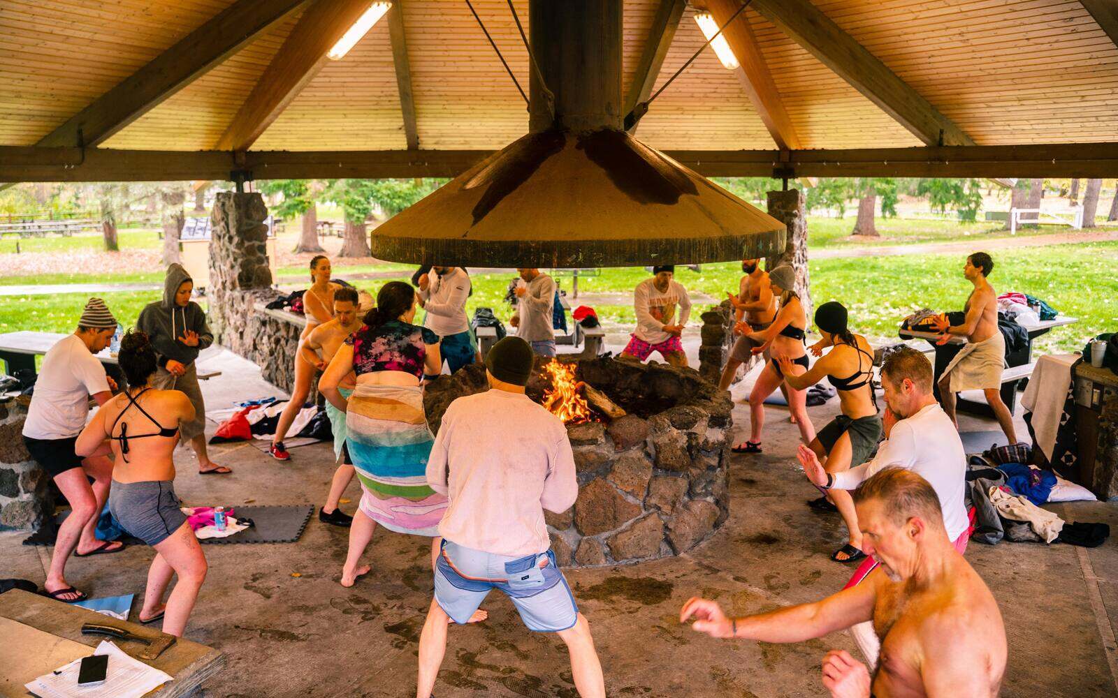 A circle of people moving and shaking around an open fire pit under a shelter